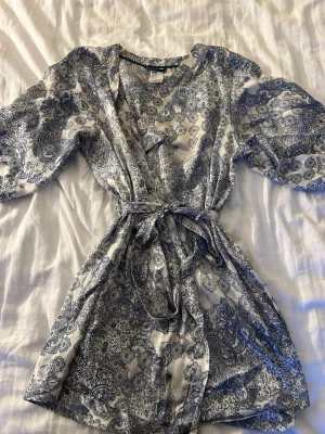 Mönstrad omlottklänning från H&M - Säljer en superfin omlottklänning från H&M i vitt och blått med vackert mönster. Klänningen har knytband i midjan, kort längd och vida ärmar. Perfekt för sommarens alla tillfällen och ger en fräsch och trendig look.