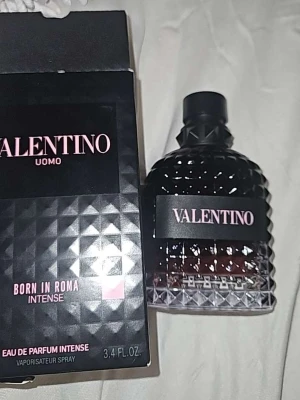 Valentino Uomo Born in Roma Intense - Säljer en lyxig parfym från Valentino, Uomo Born in Roma Intense. Flaskan är svart med en elegant mönstrad yta och rosa detaljer. Parfymen kommer i en stilren, svart kartong med rosa text. Perfekt för dig som vill ha en exklusiv doft.