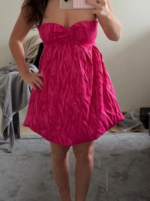 Axelbandslös rosa kortklänning - Säljer en axelbandslös kortklänning i stark rosa färg med lätt struktur i tyget. Klänningen har en söt twistad detalj vid bysten och en luftig, utställd kjol. Perfekt för dig som vill sticka ut med en färgglad och ungdomlig look.