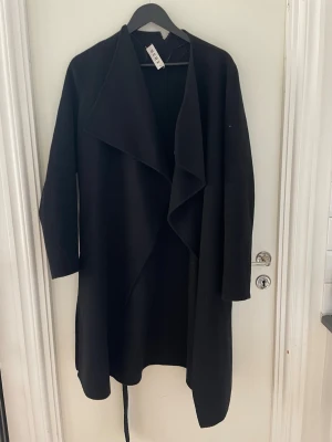 Svart kappa i ull  från WERA - Stilren svart kappa från Wera med öppen i ull front och avslappnad passform. Kappan har breda slag och är tillverkad i ett mjukt tyg som ger ett snyggt fall. Perfekt för dig som gillar minimalistisk och tidlös design.