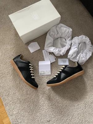 Maison Margiela Svarta Gats sneakers - Maison Margiela GATs sneakers i svart läder och mocka med beige sula och vita skosnören. Klassisk låg modell med stilren design och diskreta detaljer. Perfekta för dig som gillar minimalistisk och tidlös stil.