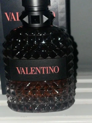 Coral fantasy - Säljer en lyxig parfym från Valentino i en mörk, nitprydd glasflaska med svart etikett och röd text. Flaskan har en unik, mönstrad yta som ger ett exklusivt intryck och känns riktigt cool att ha framme. Perfekt för dig som gillar snygg design och starka dofter.