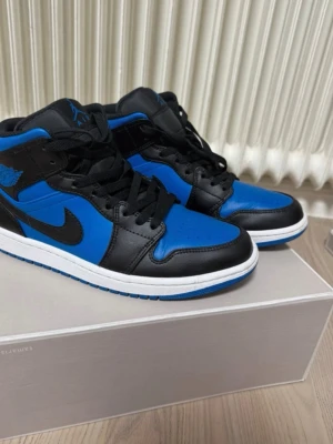 Air Jordan 1 Mid Svart/Blå - Säljer ett par Air Jordan 1 Mid i svart och blått läder med klassisk Nike swoosh på sidan. Skorna har svarta skosnören, vit mellansula och blå yttersula. Ikoniska Jordan Wings-loggan på sidan och Jumpman-logga på plösen. Perfekta för dig som gillar streetwear.