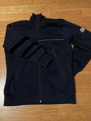 Svart zip-hoodie från Moncler - Snygg svart hoodie med dragkedja från Moncler. Har två fickor med dragkedja, ribbade muddar och Moncler-logga på ärmen. Insidan har meshfoder i orange. Perfekt för en stilren och sportig look.