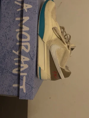 Nike Ja Morant beige/blå sneakers - Snygga Nike Ja Morant sneakers i beige och vitt med blå detaljer på sulan. Skorna har meshpaneler för extra andningsförmåga, vita swoosh-loggor på sidorna och grå/bruna partier vid hälen. Låg modell med klassisk snörning och cool signaturdetalj i rött på hälen.