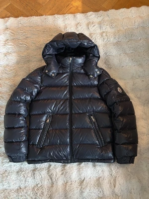 Mörkblå dunjacka från Moncler med huva - Snygg mörkblå dunjacka från Moncler med glansig finish, avtagbar huva och två dragkedjeförsedda fickor framtill. Jackan har en Moncler-logga på ärmen och quiltad design som håller värmen under kalla dagar. Den är i barnstorleken 14 år 164. Lappen med serietidningen sitter lite löst men inget som märks av (ses på bild). Den är i väldigt fint skick.  På sista bilden ser man ett äkthetsbevis som har blivit legitcheckat av checkcheck. Om ni har några funderingar så är det bara att fråga😀