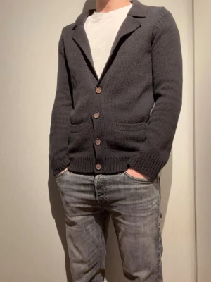 Mörkgrå stickad kofta med knappar - Snygg mörkgrå stickad kofta med fyra träknappar framtill och två fickor. Koftan har ribbade muddar vid ärmslut och nederkant samt en klassisk krage. Perfekt att slänga över en t-shirt för en avslappnad look.