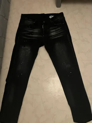 Svarta Dsquared2 jeans med slitningar - Snygga svarta jeans från Dsquared2 med coola slitningar och diskreta färgstänk. Jeansen har en smal passform och klassisk femficksdesign. Perfekta för dig som gillar streetstyle och vill sticka ut lite extra.