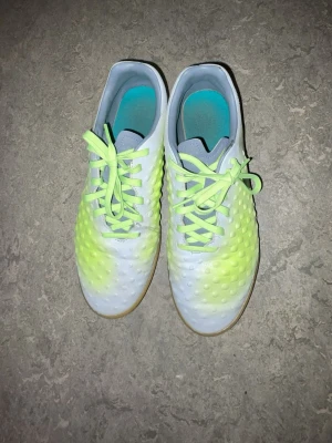 Nike vita och neongula fotbollsskor - Säljer ett par Nike fotbollsskor i vitt och neongult med svart swoosh på sidan. Skorna har en texturerad ovandel med små dobbar för bättre bollkontroll och neongröna snören. Yttersulan är i gummi, perfekt för inomhusspel.