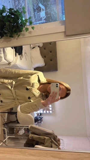 Beige trenchcoat med knytskärp - Stilren beige trenchcoat med dubbelknäppning och svarta knappar. Knytskärp i midjan och klassisk krage. Långa ärmar med slejfdetaljer vid handlederna. Perfekt för lager på lager och ger en snygg siluett.