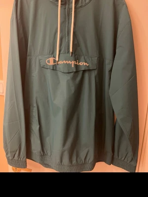 Grön vindjacka från Champion XL - Snygg grön vindjacka från Champion med vit logga på bröstet och dragkedja vid halsen. Jackan har huva med vita snören och elastiska muddar vid ärmslut och nederkant. Perfekt för blåsiga dagar och enkel att matcha med streetwear.
