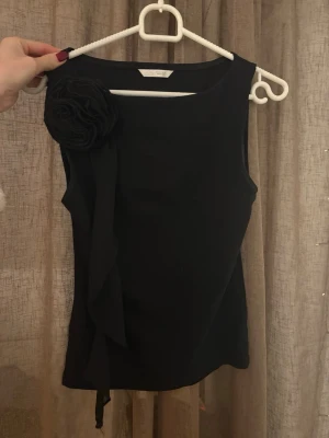 Svart ärmlös topp med blomdetalj Zara - Stilren svart ärmlös topp från Zara med en stor dekorativ blomma på axeln. Toppen har en enkel, rak passform och är tillverkad i ett mjukt material som känns bekvämt mot huden. Perfekt för dig som vill ha en snygg och unik detalj i din outfit.