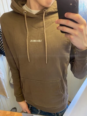 Brun hoodie från Les Deux - Snygg brun hoodie från Les Deux med vit logga på bröstet. Hoodien har huva med dragsko, känguruficka framtill och ribbade muddar vid ärmar och nederkant. Tillverkad i mjukt bomullsmaterial som är skönt att bära.