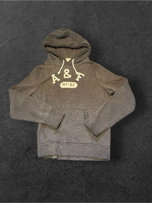 Fet Hoodie Abercrombie & Fitch - Mysig grå hoodie från Abercrombie & Fitch med broderad A&F-logga och texten NY-92 på bröstet. Klassisk känguruficka framtill och justerbar huva med vita snören. Perfekt för en avslappnad och sportig stil. 