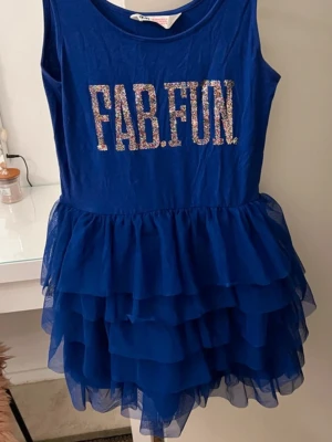 Blå volangklänning med glittertext - Snygg blå ärmlös klänning med flera lager volanger i kjolen och glittrig text 'FAB.FUN' framtill. Klänningen har rund halsringning och är tillverkad i ett lätt, luftigt material som ger en festlig känsla.