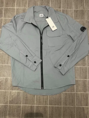 Ljusblå overshirt från C.P. Company - Snygg ljusblå/grå overshirt från C.P. Company med svart dragkedja, bröstficka och knappdetaljer på ärmarna. Klassisk krage och rundad nederkant. Tillverkad i ett lätt och slitstarkt material, perfekt för lager på lager. Ikonisk logga på ärmen.