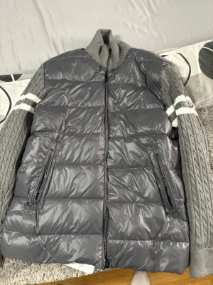 Grå Moncler dunväst med stickade ärmar - Snygg grå dunväst från Moncler med glansig framsida och stickade ärmar i kabelstickat mönster. Två dragkedjefickor fram och vita ränder på ärmarna. Hög krage och Moncler-logga på vänster ärm. Perfekt för lager-på-lager och streetstyle. Passar även om du har storlek L, pga att den är liten i storleken. Vid snabbaffär kan priset sänkas!