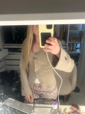 Beige kofta med vida ärmar - Säljer en beige kofta med stora knappar och vida ärmar. Koftan har v-ringning och dekorativa knappar vid ärmsluten. Perfekt för dig som gillar en avslappnad men ändå trendig stil. Materialet känns mjukt och koftan har en lite boxig passform.