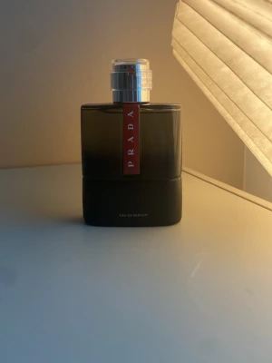 Prada Eau de Parfum carbon 100 ml - Snygg parfym från Prada i en stilren svart glasflaska med silverfärgad kork och röd etikett framtill. Flaskan har en modern, fyrkantig form och känns lyxig och elegant. Perfekt för dig som gillar exklusiva dofter och vill ha en parfym som sticker ut i badrumshyllan.