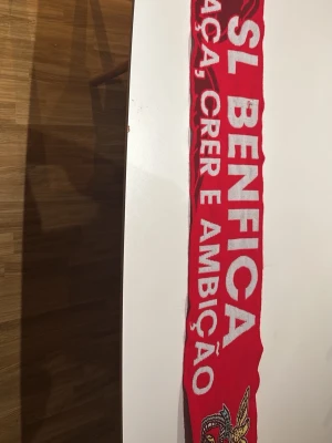 Röd SL Benfica halsduk - Röd halsduk med SL Benfica-logga och texten 'SL BENFICA' i vitt. Halsduken har fransar i ändarna och detaljer i gult, svart och grönt. Perfekt för dig som vill visa ditt stöd för Benfica. Materialet är stickat och mjukt.