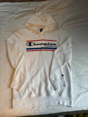 Vit hoodie från Champion - Vit hoodie från Champion med stor logga och texten 'New York EST. 1919' på bröstet. Hoodien har röda och blå detaljer, känguruficka framtill och snöre i huvan.
