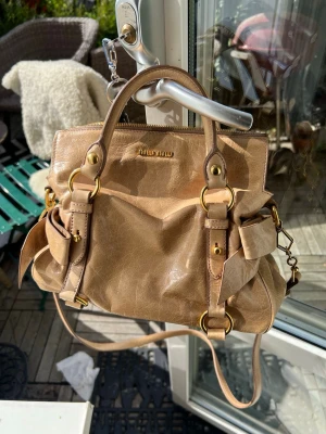 Beige handväska från Miu Miu - Säljer en snygg beige handväska från Miu Miu i glansigt läder. Väskan har guldfärgade metalldetaljer, dubbla handtag och justerbar axelrem. Flera fickor på sidorna och coola spännen ger den en trendig look. Perfekt för dig som gillar lyxiga accessoarer.