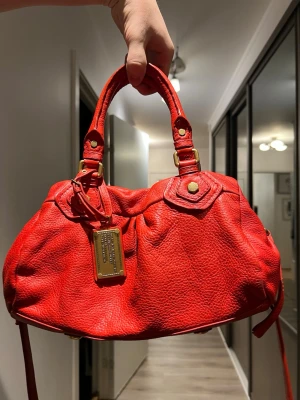 Röd handväska från Marc by Marc Jacobs - Säljer en snygg röd handväska från Marc by Marc Jacobs i mjukt skinn. Väskan har guldfärgade metalldetaljer, dubbla handtag och en axelrem. Märkets klassiska metallbricka framtill ger en lyxig touch. Perfekt för dig som vill sticka ut med färg!