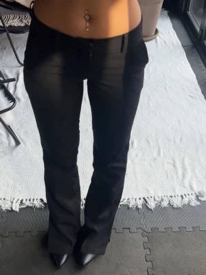 Svarta lågmidjade kostymbyxor - Aldrig använda! Snygga svarta kostymbyxor med låg midja och utsvängda ben. Byxorna har hällor för skärp, två fickor fram och två dekorativa fickor bak. Perfekta för en trendig och stilren look.