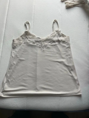 Vit spetsdetaljerad linnetopp - Säljer en stilren vit linnetopp med tunna axelband och vacker spets längs urringningen. Toppen har en mjuk och lätt känsla, perfekt för lager på lager eller sommarens varma dagar. Enkel att matcha med både jeans och kjol. Har några fläckar 