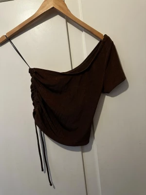 Brun oneshoulder topp från H&M Divided - Säljer en brun oneshoulder topp från H&M Divided. Toppen har kort ärm, snörning på sidan och en croppad passform. Perfekt för dig som vill ha en trendig och unik look med snygga detaljer. Materialet känns mjukt och stretchigt.