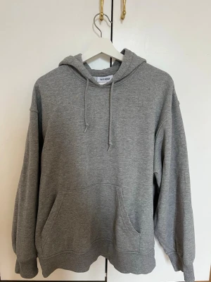 Grå hoodie från Weekday - Klassisk oversized grå hoodie från Weekday med huva och dragsnören. Hoodien har en stor magficka framtill och ribbade muddar vid ärmar och nederkant. Storlek Xs