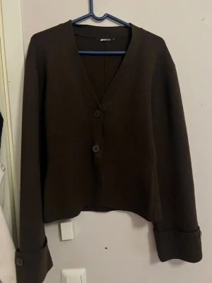 Mörkbrun kofta från Gina Tricot - Stilren mörkbrun kofta från Gina Tricot med v-ringning och två stora knappar framtill. Koftan har långa ärmar med breda muddar och en rak, något kortare passform. Perfekt för lager på lager och enkel att matcha.