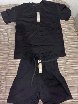 Essentials Fear of God set t shirt och shorts - Snyggt svart set från Essentials Fear of God med t-shirt och shorts. Setet har diskret logga på shortsen och är gjort i mjukt bomullsmaterial. Perfekt för en avslappnad och trendig look. T-shirten har rund hals och shortsen har dragsko i midjan.