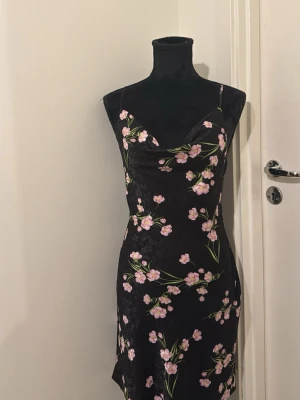 Svart blommig klänning med smala band 🩷💚 - Supersnygg svart klänning med smala axelband och blommigt mönster i rosa och grönt. Klänningen har en figurnära passform och lätt glansigt material som ger en lyxig känsla. Perfekt för dig som vill sticka ut med en feminin och trendig look.