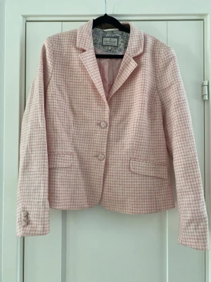 Rosa och vit kavaj från Newhouse - Snygg kavaj i rosa och vitt hundtandsmönster från Newhouse. Klassisk modell med två knappar framtill, fickor och tre dekorativa knappar vid ärmslutet. Tunt fodrad och i ett mjukt tyg som känns lyxigt. Perfekt för dig som vill ha en preppy och trendig look. Ny, aldrig använd  Mått Över bröstet: ca 53 cm Längd från axeln och ner: ca 59 cm  100% ull  Kommer från ett djur och rökfritt hem