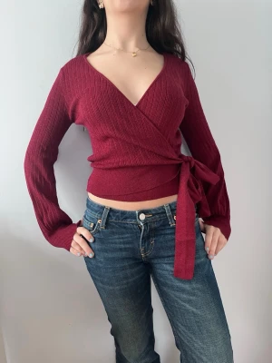 Vinröd omlottopp med knytband - Trendig vinröd och glittrig omlottopp med långa ärmar och breda ribbade muddar. Väldig söt och skön, nästan helt oanvänd!💕