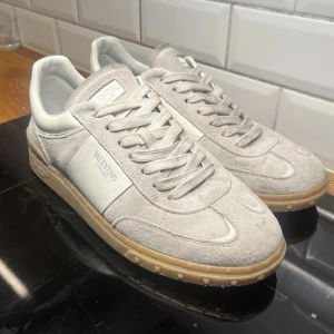 Valentino ljusgrå sneakers i mocka - Snygga ljusgrå sneakers från Valentino med ovandel i mjuk mocka och vita detaljer på sidan. Skorna har en klassisk låg siluett, vita skosnören och en beige gummisula som ger en clean look. Perfekta för dig som gillar stilrena och exklusiva sneakers.