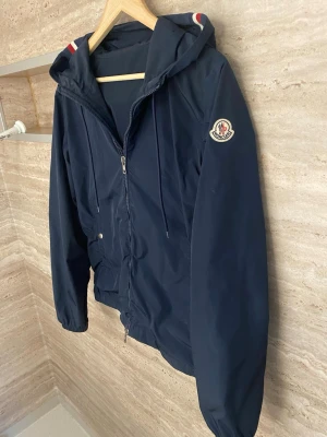 Mörkblå vindjacka från Moncler - Snygg mörkblå vindjacka från Moncler med huva och dragkedja framtill. Jackan har två fickor med tryckknappar och Moncler-logga på ärmen. Detaljer med röd och vit rand på huvan ger en sportig touch. Tillverkad i lätt och tåligt material, perfekt för blåsiga dagar.skriv vid frågor
