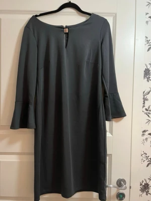 Mörkgrå klänning med trumpetärm - Stilren mörkgrå klänning med långa trumpetärmar och rund halsringning. Klänningen har en liten slits framtill vid halsen och dragkedja bak. Rak passform och enkel design som passar till många tillfällen.