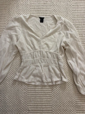 Vit blus med v-ringning från Vero Moda - Säljer en vit blus från Vero Moda med v-ringning och långa puffärmar. Blusen har en lätt rynkad struktur i tyget och smock i midjan för en snygg passform. Tillverkad i ett tunt, luftigt material som känns skönt mot huden.