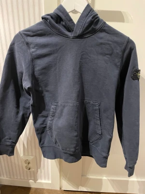 Mörkblå hoodie från Stone Island - Snygg mörkblå hoodie från Stone Island med klassisk patch på ärmen. Hoodien har en stor känguruficka framtill och en skön huva. Perfekt för dig som gillar streetwear och vill ha något stilrent men ändå med edge.