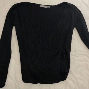 Svart omlottkofta från KappAhl - Svart ribbad omlottkofta från KappAhl i storlek M. Koftan har långa ärmar, knyt i sidan och djup v-ringning. Perfekt att slänga över ett linne eller t-shirt för en chill och snygg look. Materialet är mjukt och stretchigt, vilket gör den riktigt skön att bära.❣️❣️