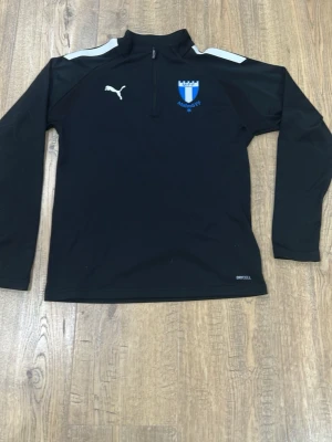 Svart Malmö FF träningströja Puma - Svart långärmad träningströja från Puma med Malmö FF-logga på bröstet. Tröjan har halv dragkedja, vita detaljer på axlarna och är tillverkad i funktionsmaterial med DryCell-teknologi. Perfekt för fotbollsträning eller sportaktiviteter.