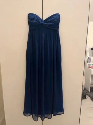 Mörkblå axelbandslös balklänning - Säljer en mörkblå axelbandslös långklänning med twistad överdel och plisserad kjol. Klänningen har en elegant, flowy passform och är gjord i ett tunt, svepande tyg som ger en lyxig känsla. Perfekt till balen eller en superfin strandklänning!💕