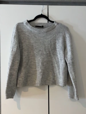 Ljusgrå stickad tröja - Mysig ljusgrå stickad tröja med rund halsringning och långa ärmar. Tröjan har en enkel, rak passform och ribbstickade muddar vid hals, ärmslut och nederkant. Perfekt för lager på lager under kyliga dagar.