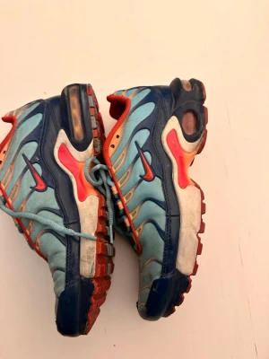 Nike Air Max Plus blå och orange - Nike Air Max Plus sneakers i blått och ljusblått med orange detaljer och röd swoosh. Ovandelen är i mesh och syntet, med vågiga linjer och synlig Air Max-dämpning i sulan. Insidan är orange och sulan har inslag av rött och blått. Klassisk streetwear-look.