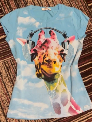 Blå t-shirt med giraff och hörlurar - Cool blå t-shirt med molnigt mönster och en färgglad giraff med hörlurar och hjärthalsband på framsidan. T-shirten har korta ärmar och figurnära passform. Perfekt för dig som gillar unika och lekfulla plagg.