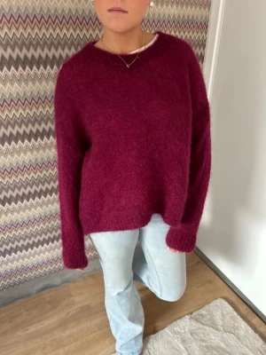 Vinröd fluffig stickad tröja - Mysig vinröd stickad tröja med oversized passform och långa ärmar. Tröjan har en enkel rund halsringning och är gjord i ett fluffigt material som ger en trendig look. Perfekt att matcha med jeans för en avslappnad stil.