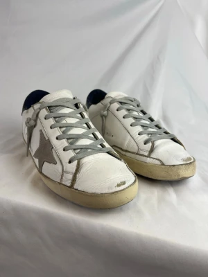 Golden Goose Superstar Sneakers  - Säljer ett par vita Golden Goose sneakers med grå stjärna på sidan och grå skosnören. Skorna har en klassisk låg siluett och blå detalj på hälen. Ovandelen är i läder och sulan är något gulnad för en vintage look.