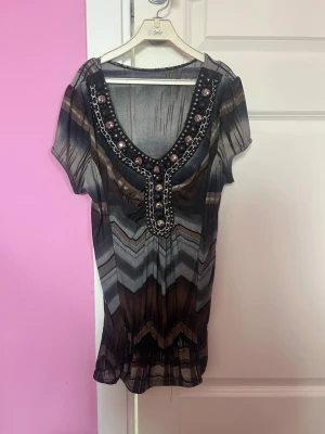 Mönstrad topp med kedjedetaljer - Säljer en unik topp med zigzagmönster i svart, grått och brunt. Toppen har korta ärmar och en djup v-ringning dekorerad med kedjor och pärlor. Materialet är tunt och lätt, perfekt för dig som gillar statement-detaljer.
